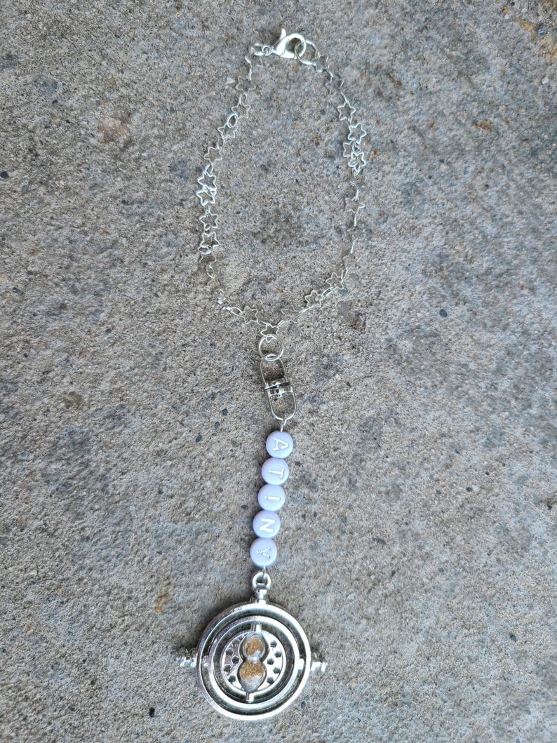 May include: A silver charm necklace with a star-shaped chain and a silver hourglass charm. The charm has a white beaded chain with the word "ATINE" spelled out in white beads.