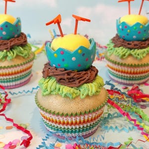 Könnte beinhalten: Drei dekorierte Cupcakes mit gelbem, blauem und braunem Zuckerguss, garniert mit orangefarbenen Elementen. Die Cupcakes stehen auf einer Oberfläche mit bunten Konfetti und dem Text "Imagine Out Loud". Die Cupcakes haben ein festliches Aussehen.