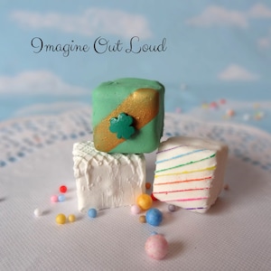 May include: Three decorative, square-shaped treats are stacked on a white doily. The top treat is green with a gold stripe and a shamrock. The middle treat is white with a textured design. The bottom treat has rainbow stripes. The text "Imagine Out Loud" is in the background.
