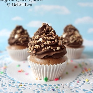 Puede incluir: Tres cupcakes de chocolate con glaseado marrón y chispas sobre un encaje blanco. Los cupcakes están decorados con virutas de chocolate y chispas blancas. El texto "Imagine Out Loud © Debra Lea" es visible en el fondo.