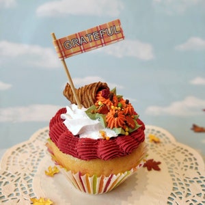 May include: A red and white cupcake with a cornucopia and fall-themed decorations. The cupcake is topped with white frosting and a small flag that says "GRATEFUL". The cupcake is sitting on a white doily.