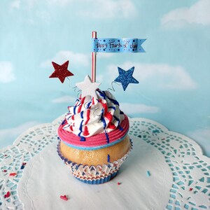 May include: A red, white, and blue cupcake with a white frosting swirl and a blue and red striped design. The cupcake is topped with a white star and a blue flag that says "Happy Fourth of July".