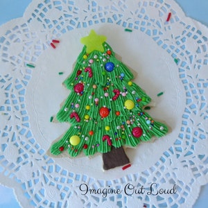 Può includere: Un biscotto a forma di albero di Natale glassato di verde decorato con codette colorate, caramelle e una stella gialla in cima. Il biscotto è su un tovagliolo di pizzo bianco.