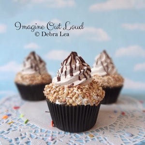 Könnte beinhalten: Drei Cupcakes mit weißem Zuckerguss, gehackten Nüssen und Schokoladenstreuseln werden auf einer weißen Spitze präsentiert. Die Cupcakes befinden sich in schwarzen Papierförmchen. Das Bild enthält den Text "Imagine Out Loud" und "© Debra Lea".