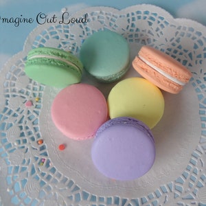 May include: Five pastel-colored macarons on a white doily. The macarons are green, blue, pink, yellow, and purple.