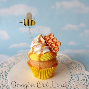 Puede incluir: Un cupcake amarillo con glaseado blanco en espiral, coronado con un panal de abejas y una pequeña figura de abeja. El cupcake está sobre un encaje blanco con el texto "Imagine Out Loud" debajo.