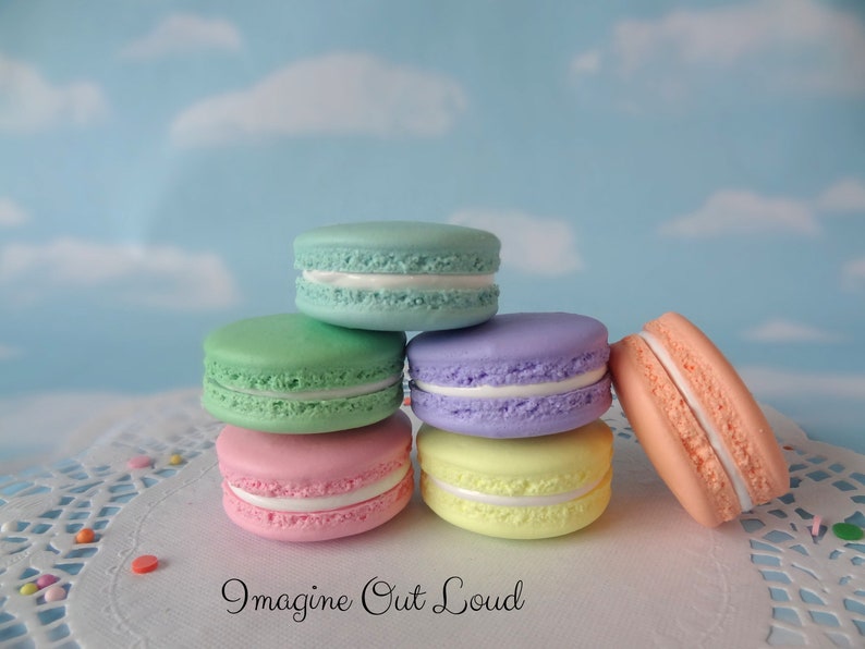 May include: Six pastel-colored macarons stacked on a white doily with white frosting and a light blue background.  The macarons are in the following order from top to bottom: light blue, green, purple, pink, yellow, and peach.