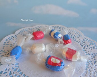 May include: Six wrapped candies in red, white, and blue colors on a white doily. The candies are arranged in a circle. The background is a light blue sky with white clouds.