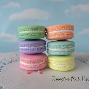 May include: A stack of six pastel-colored macarons with white filling. The macarons are arranged in a pyramid shape on a white doily. The background is a light blue sky with white clouds.