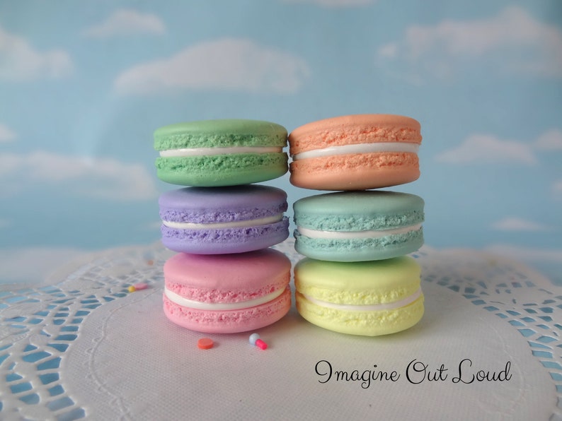 May include: A stack of six pastel-colored macarons with white filling. The macarons are arranged in a pyramid shape on a white doily. The background is a light blue sky with white clouds.