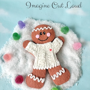 Può includere: Un biscotto a forma di omino di pan di zenzero con un maglione bianco con un cuore rosso, circondato da caramelle colorate e neve artificiale. Il testo "Imagine Out Loud" è scritto in corsivo sopra il biscotto su sfondo azzurro.