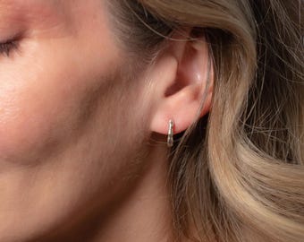Op de afbeelding: Een close-up van een oor met een kleine, zilveren oorring. De oorring heeft een textuur en wordt in de oorlel gedragen. De persoon heeft blond haar en een lichte huid.