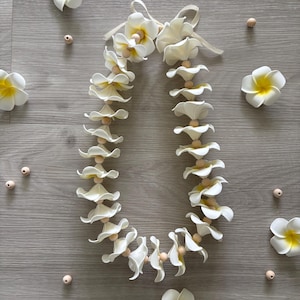 Könnte beinhalten: Eine weiße Lei-Halskette mit cremefarbenen Blüten und Holzperlen. Mehrere lose Blüten und Perlen sind um die Lei auf einer grauen Holzoberfläche verstreut. Die Blüten haben gelbe Zentren.