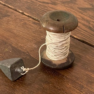 May include: A wooden spool of white string with a plumb bob attached. The spool is made of dark wood and has a hole in the center. The plumb bob is a gray, pyramid-shaped weight.