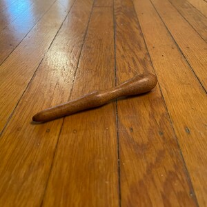 May include: A handcrafted wooden massage tool with a smooth, ergonomic design. The tool is a warm brown color and appears to be made from a single piece of wood. It is lying on a wooden floor.