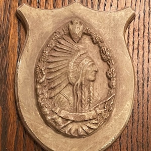 May include: A brown, shield-shaped plaque with a raised relief sculpture of a Native American man wearing a feathered headdress. The sculpture is surrounded by a wreath of leaves and flowers.