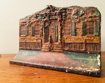 May include: A vintage cast iron doorstop shaped like a house with a brown, yellow, and green painted exterior. The house has a front porch with a railing and a small fence on either side. The house has two windows on each side and a front door with a small window.