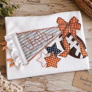 May include: A white t-shirt featuring an applique design of a gray striped pennant with the word "Jols" embroidered in orange, a brown football with white laces, an orange gingham bow, and three stars in white, orange gingham, and navy blue.