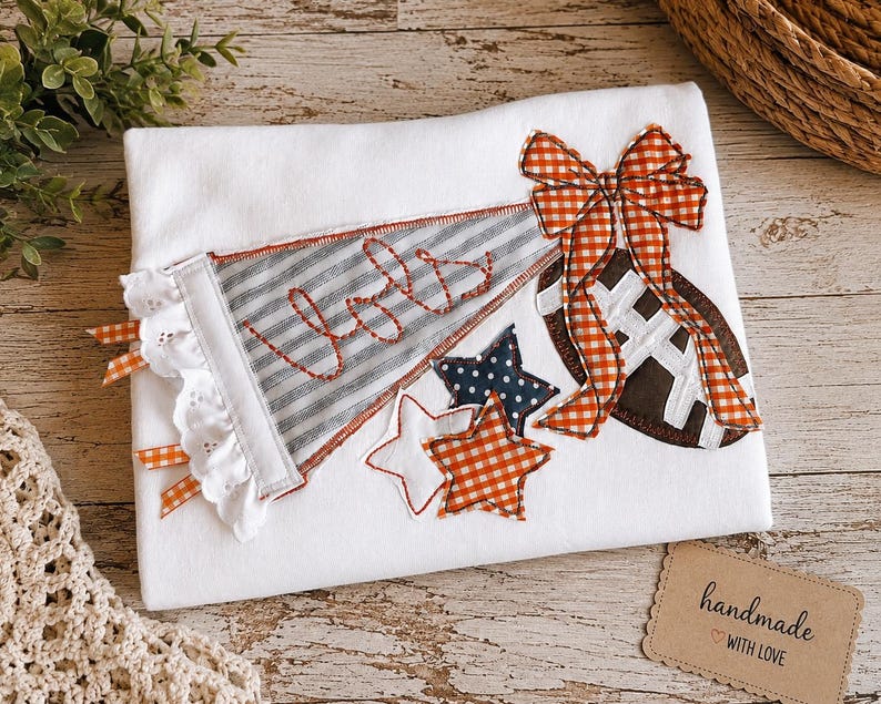 May include: A white t-shirt featuring a fabric applique design of a gray striped pennant with the word "Jols" embroidered in red, a brown football with white laces, an orange gingham bow, and three stars in white, orange gingham, and navy blue.