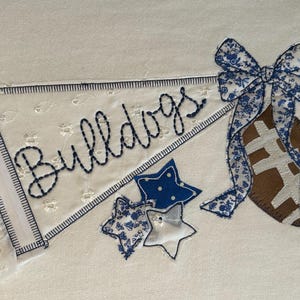 May include: An embroidered pennant on white fabric featuring the word Bulldogs in dark blue stitching. The design includes a brown football with a blue and white floral bow, three star-shaped fabric appliques, and blue gingham ribbons.