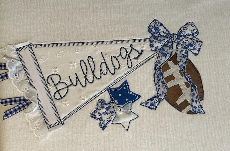 May include: An embroidered pennant on white fabric featuring the word Bulldogs in dark blue stitching. The design includes a brown football with a blue and white floral bow, three star-shaped fabric appliques, and blue gingham ribbons.