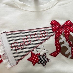 May include: A white sweatshirt featuring an appliqued pennant with black and white stripes and the red embroidered text Roll Tide. The design includes a brown football, a red and white polka dot bow, and three small stars in red, white, and black.