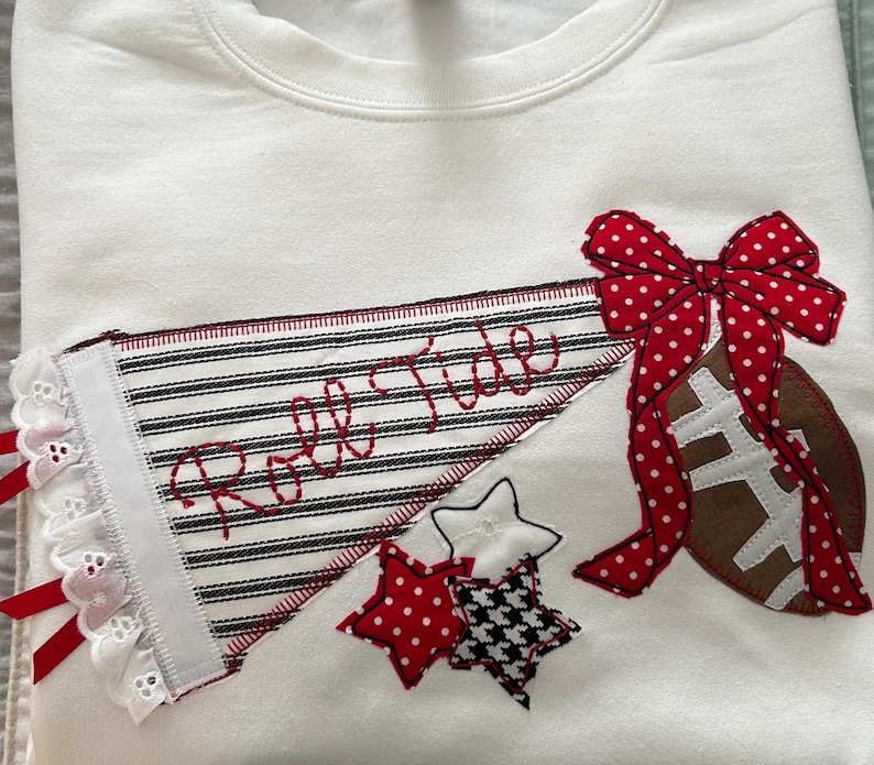 May include: A white sweatshirt featuring an appliqued pennant with black and white stripes and the red embroidered text Roll Tide. The design includes a brown football, a red and white polka dot bow, and three small stars in red, white, and black.