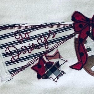 May include: A white fabric applique featuring a striped pennant with the embroidered text Go Daddy, a brown football, a red polka dot bow, and a plaid star. The pennant is accented with white eyelet lace trim and black ribbon details.