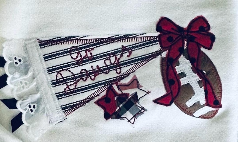 May include: A white fabric applique featuring a striped pennant with the embroidered text Go Daddy, a brown football, a red polka dot bow, and a plaid star. The pennant is accented with white eyelet lace trim and black ribbon details.