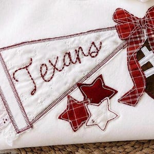 May include: A white fabric item featuring an embroidered pennant with the word Texans in red stitching, a brown football with white laces, a red and white plaid bow, and three small stars in red and white fabric appliqued onto the surface.