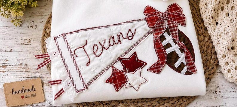 May include: A white fabric item featuring an embroidered pennant with the word Texans in red stitching, a brown football with white laces, a red and white plaid bow, and three small stars in red and white fabric appliqued onto the surface.
