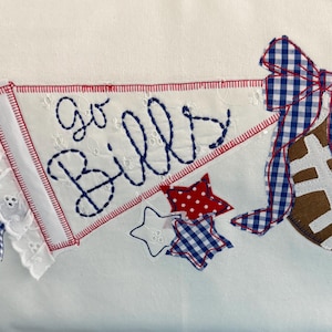 May include: A white fabric pennant with eyelet lace trim and blue stitched text reading Go Bills. A brown football is attached to the right side, adorned with a blue and white gingham ribbon bow. Two small stars, one red with white dots and one blue gingham, sit below.