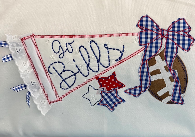 May include: A white fabric pennant with eyelet lace trim and blue stitched text reading Go Bills. A brown football is attached to the right side, adorned with a blue and white gingham ribbon bow. Two small stars, one red with white dots and one blue gingham, sit below.