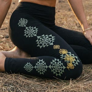 May include: A black racerback crop top featuring a gold circular geometric mandala print on the chest, paired with black leggings that display a vertical row of repeating triangular geometric patterns in shades of green and gold along the side.