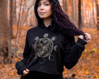 May include: A black cropped hoodie with a gold moon and flower of life design. The hoodie has a drawstring hood and long sleeves.