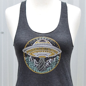 May include: Dark gray tank top with a round graphic of a UFO over a mountain scene. The UFO is silver and gold, with a yellow and green border. The tank top is displayed on a mannequin.