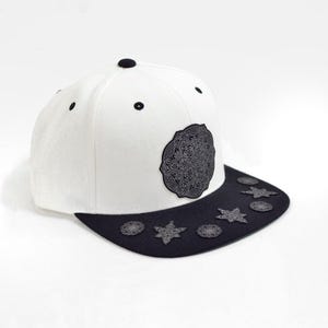May include: A white baseball cap with a black flat brim. The front panel features a circular black patch with an intricate mandala-style design. The black brim is decorated with several small, dark, star-shaped and circular metallic-looking studs.