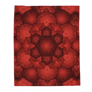 May include: A red blanket with a detailed, symmetrical pattern. The design features repeating floral and geometric shapes in varying shades of red, creating a layered effect. The blanket is rectangular and appears to be made of soft fabric.