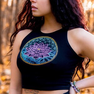 May include: A woman with long dark hair wearing a black crop top with a colorful lotus flower design. The top has a halter neck style and is cropped above the waist. The woman is standing in a forest setting with fall foliage on the ground.