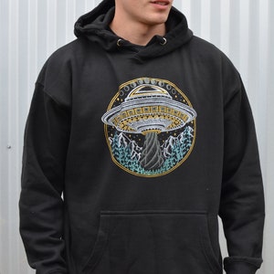 May include: Black hooded sweatshirt with a large, circular graphic of a UFO hovering over a mountain landscape. The UFO is detailed with gold and white accents, and the mountains are depicted in shades of blue and green.