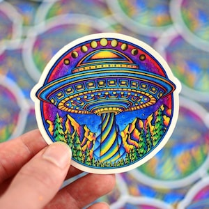 Puede incluir: Una pegatina colorida con un OVNI psicodélico flotando sobre una cordillera con un fondo de cielo estrellado. El OVNI tiene una cúpula amarilla y naranja con un cuerpo azul y morado. La cordillera es una mezcla vibrante de azules, verdes y amarillos. La pegatina es circular y tiene un borde blanco.