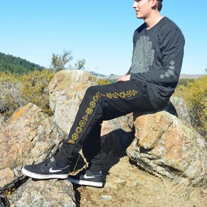 May include: A black baseball cap with a graphic, a charcoal gray long-sleeve shirt featuring a large geometric print on the chest and sleeve, and black joggers with a vertical row of yellow geometric shapes along the side, paired with black sneakers.