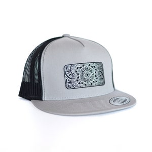 May include: A grey and black trucker hat with a black mesh back. The front of the hat features a silver rectangular patch with a detailed black and white geometric design.