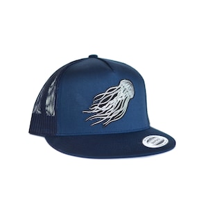 Puede incluir: Una gorra de camionero azul marino con un gráfico de medusa blanco y negro. La gorra tiene una parte trasera de malla y una visera curvada.