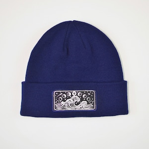 May include: A navy blue beanie with a black and white embroidered patch featuring a wave and a crescent moon with stars.