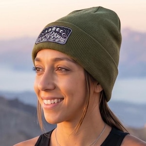 May include: Black tank top with a circular mountain and moon graphic. The woman is wearing an olive green beanie with a mountain patch, olive green leggings, and an orange climbing harness. The background shows a mountain range.