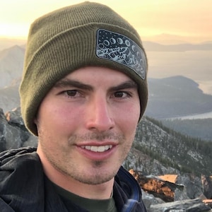 Pode incluir: Uma pessoa usando um gorro verde-oliva com um patch com um design de fases da lua. O fundo mostra uma paisagem de montanhas e lagos, com um céu laranja e amarelo ao amanhecer.