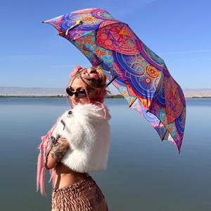 May include: A vibrant umbrella featuring a complex, multi-colored mandala pattern in shades of blue, purple, pink, and orange. The canopy displays intricate geometric and circular designs that radiate from the center, creating a bold visual display.