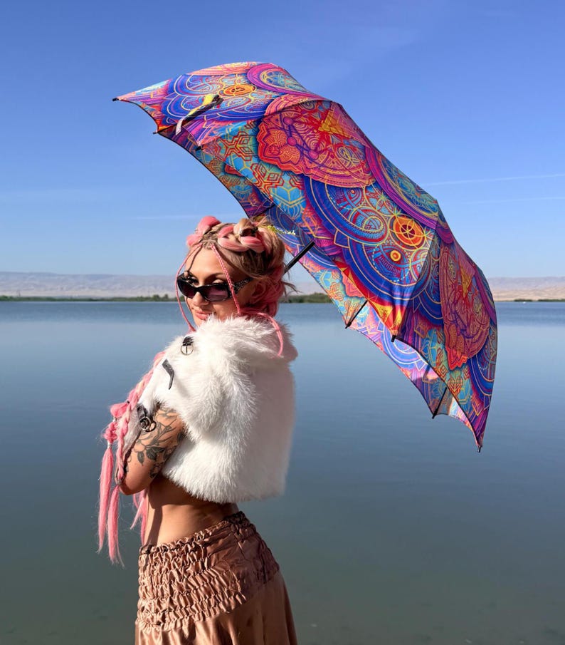 May include: A vibrant umbrella featuring a complex, multi-colored mandala pattern in shades of blue, purple, pink, and orange. The canopy displays intricate geometric and circular designs that radiate from the center, creating a bold visual display.