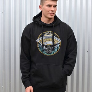 May include: Black hooded sweatshirt with a large circular graphic of a UFO, mountains, and trees. The graphic is in yellow, white, and blue. The sweatshirt has a front pocket and a drawstring hood.